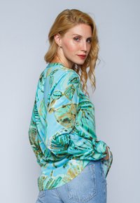 Langärmlige Bluse aus glänzendem, hellblauem Stoff mit einem abstrakten Blattmuster in Grün und Braun. Lockere Passform mit gerundetem Saum, leicht eingesteckt.