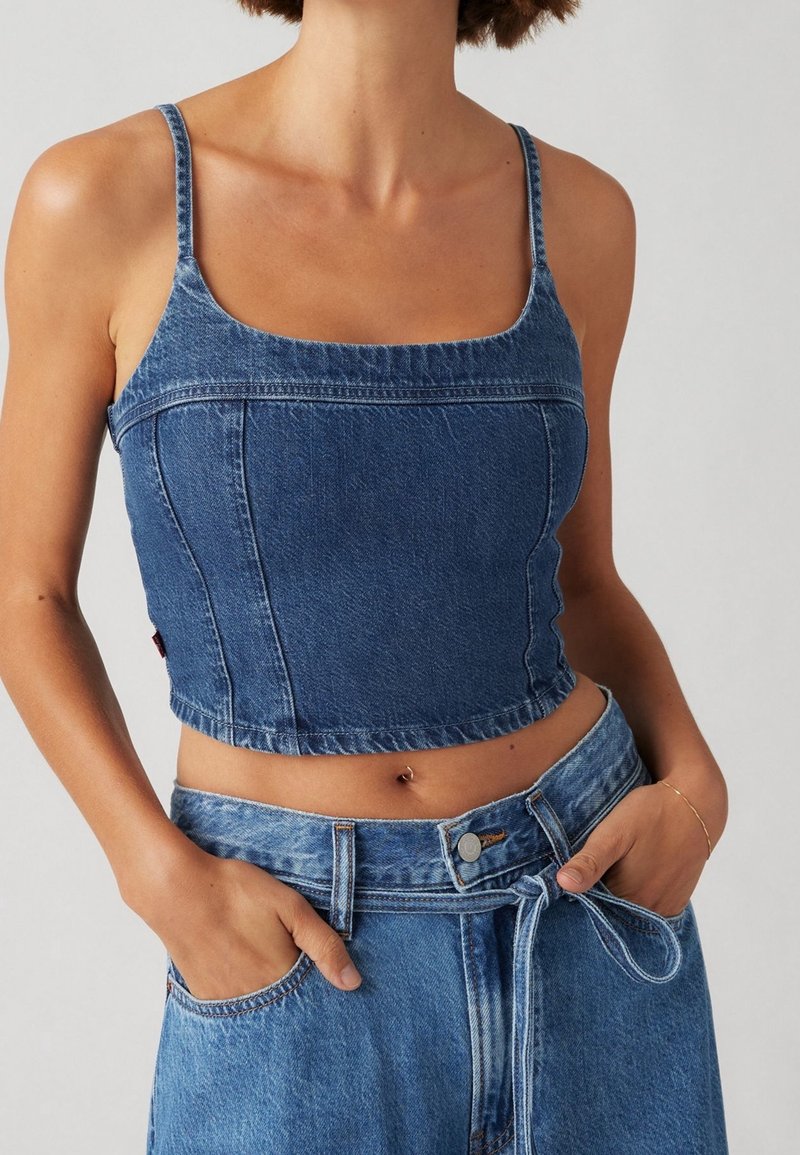 Femme portant un crop top en jean sans manches et un jean taille haute avec une ceinture nouée, les mains dans les poches, sur un fond neutre.
