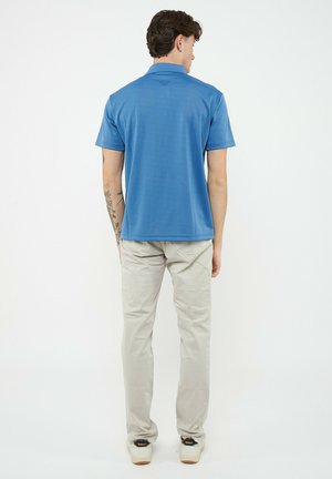 Uomo con braccio sinistro tatuato che indossa una polo blu a maniche corte, pantaloni beige e scarpe da ginnastica bianche, in piedi di schiena su sfondo bianco.