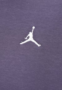 Tela morada con un logo Jumpman bordado en blanco, que representa a una persona en el aire, alcanzando una pelota de baloncesto. Textura suave.