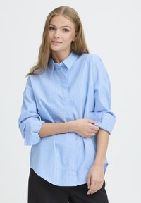 Svetlo modra srajca s gumbom in belimi navpičnimi črtami, z zvitimi rokavi in sproščenim krojem. Material izgleda gladek in lahek.