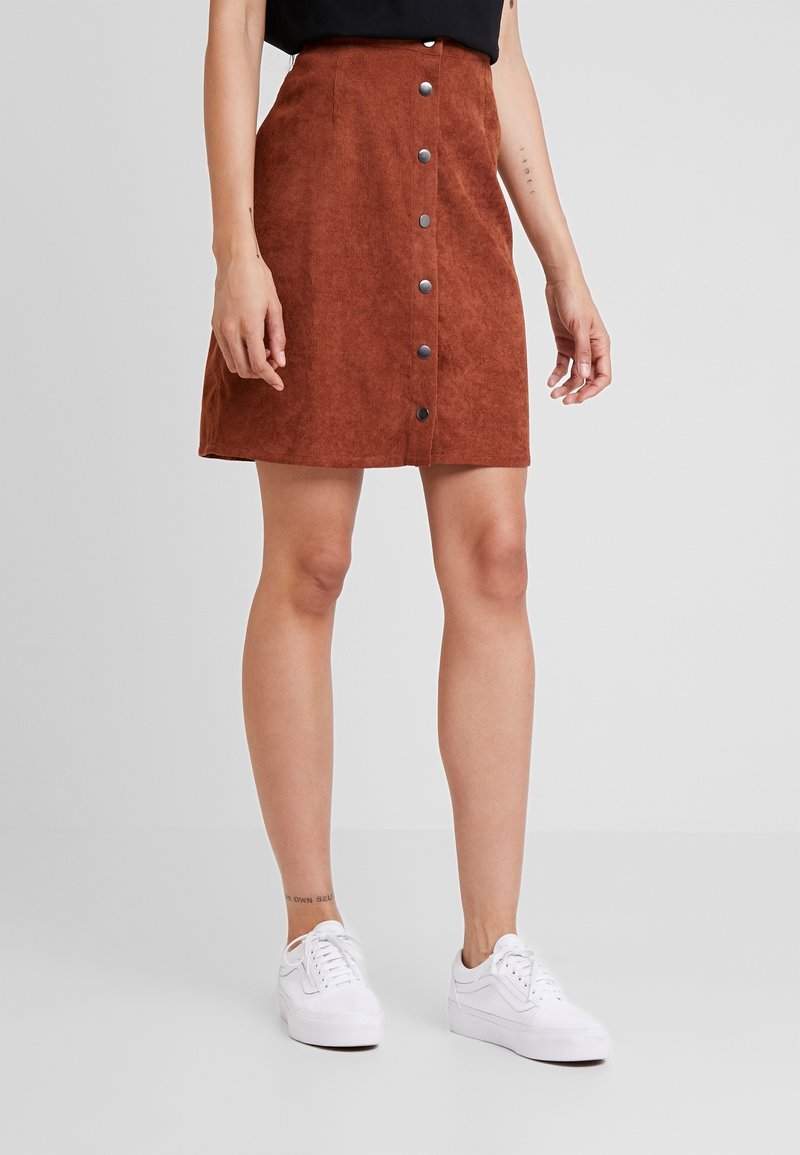 Femme portant une jupe en velours côtelé couleur rouille boutonnée devant, avec des baskets blanches, debout sur un fond gris uni.