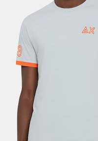 Maglietta grigia chiara a maniche corte con un accento arancione sulla manica, caratterizzata da ricami geometrici arancioni. Texture liscia.