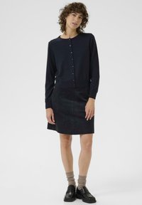 Marineblauer Cardigan mit Knopfleiste und geripptem Saum, kombiniert mit einem schwarzen Baumwollrock. Das Model trägt schwarze Loafers und strukturierte beige Socken.