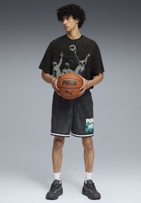 Camiseta gráfica negra con diseño de baloncesto, pantalones cortos de malla negros con acentos en blanco y verde azulado, sosteniendo un balón de baloncesto marrón, llevando zapatillas grises.