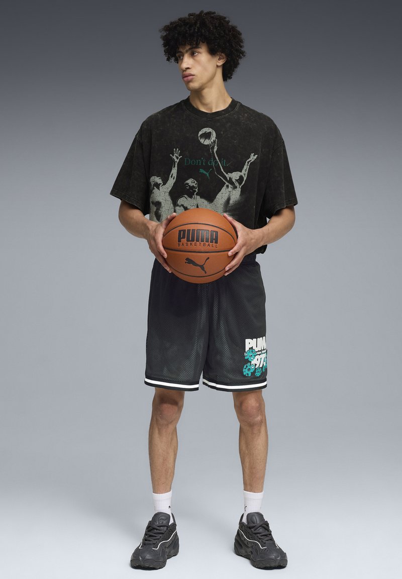 Camiseta gráfica negra con diseño de baloncesto, pantalones cortos de malla negros con acentos en blanco y verde azulado, sosteniendo un balón de baloncesto marrón, llevando zapatillas grises.
