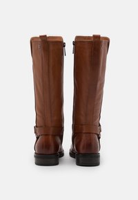 Botas de cuero marrón hasta la rodilla con cremalleras laterales, correas decorativas y un tacón apilado. La textura es suave con detalles de costura sutiles.