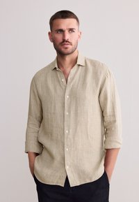 Camicia beige chiaro a righe con bottoni, vestibilità comoda, maniche lunghe arrotolate fino ai gomiti e bottoni bianchi, abbinata a pantaloni scuri.