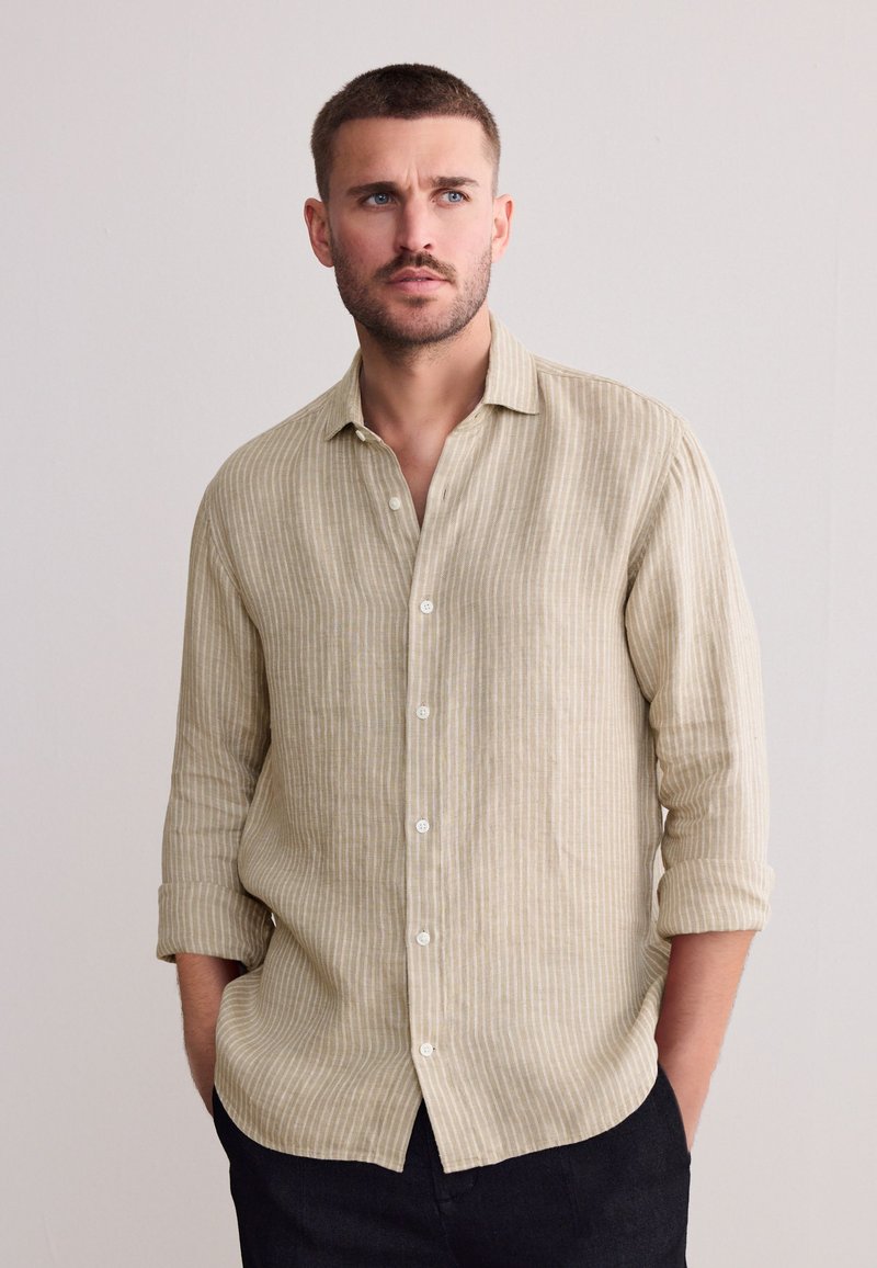 Camicia beige chiaro a righe con bottoni, vestibilità comoda, maniche lunghe arrotolate fino ai gomiti e bottoni bianchi, abbinata a pantaloni scuri.