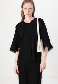 Black top with fringe detailing at sleeves and neckline, featuring a loose fit and textured fabric. Paired with a white mini bag.