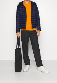 Marinblå puffjacka, orange tröja, svarta byxor och vita skor. Håller en svart toteväska. Modern design med kontrasterande färger och texturer.