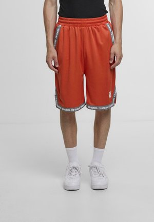 Muž oblečený v oranžových športových šortkách po kolená s bielym lemovaním a bielych teniskách na neutrálnom pozadí.