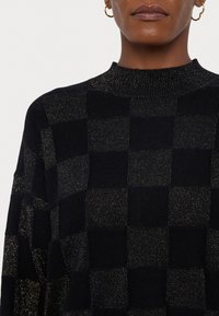 Pull en tricot noir avec un motif à carreaux et des accents métalliques dorés, col haut et tissu texturé.