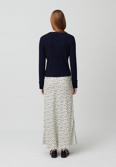 Pull côtelé bleu marine à manches longues, associé à une jupe blanche avec un petit motif à pois noirs, et des chaussures noires.