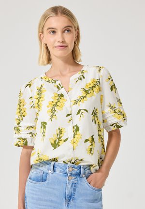 Femme aux cheveux blonds portant un chemisier blanc avec un imprimé floral jaune et un jean bleu clair, debout avec une main dans la poche.