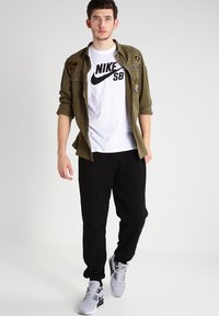 Zaļš pogājams krekls ar plāksteriem, balts Nike SB t-krekls, melni sporta bikses un pelēki čības, kas demonstrē ikdienišķo ielu modes stilu.