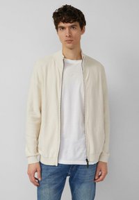 Lichtbeige vest met ritssluiting, geribbelde kraag en manchetten, textuurrijke gebreide stof, gedragen over een wit T-shirt en gecombineerd met een blauwe spijkerbroek.