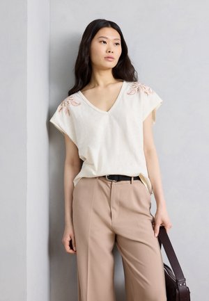 Νεαρή γυναίκα με μακριά σκούρα μαλλιά, φοράει εκρού κεντημένη μπλούζα με ντεκολτέ σε σχήμα V και μπεζ παντελόνι, κρατώντας μια σκουρόχρωμη καφέ τσάντα, στέκεται μπροστά σε έναν γκρι τοίχο.