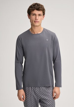 Dlouhý rukáv šedá košile vyrobená z měkké tkaniny, s kulatým výstřihem a malým, jednoduchým logem na hrudi. V kombinaci s vzorovanými kalhotami.