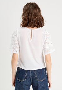 Weißes kurzärmeliges Oberteil mit Spitzenärmeln im Blumenmuster und einer Knopfverschluss hinten; getragen mit blauen Jeans.