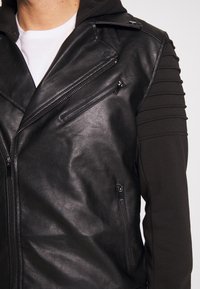 Giubbotto in pelle nera con chiusura a zip frontale, accentuazioni sulle spalle e cappuccio. Presenta pannelli testurizzati e diverse tasche con zip.