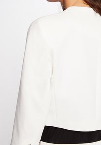Vit skräddarsydd jacka med rundade halslinjer och långa ärmar, med en svart panel längst ner. Slät textur och strukturerad design.