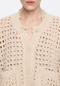 Cardigan lavorato a maglia in beige chiaro con design a trama aperta, dotato di scollo rotondo, chiusura frontale con bottoni e due tasche.