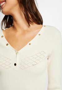 Weißer, gerippter Pullover mit V-Ausschnitt, dekorativen goldenen Knöpfen und diamantförmigen Spitzenmustern auf den Schultern. Strukturiertes Gewebe.