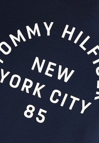 Bílý text "Tommy Hilfiger New York City 85" vytištěný na tmavě modrém textilním pozadí.