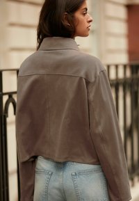 Brun ruskind cropped jakke med en blød tekstur, der har en klassisk krave og minimal syning. Båret over lyseblå denimjeans.