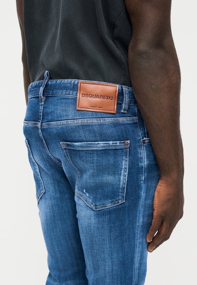 Homme portant un jean bleu usé Dsquared2 et un t-shirt noir, vu de dos de la taille à la mi-cuisse, main reposant le long du corps.