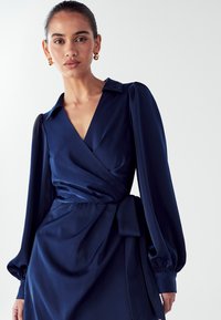 Námornícky modré zavinovacie šaty z saténového materiálu, s límcom s výrezom, dlhými vypuklými rukávmi a detailom v podobe mašle na boku v oblasti pása.