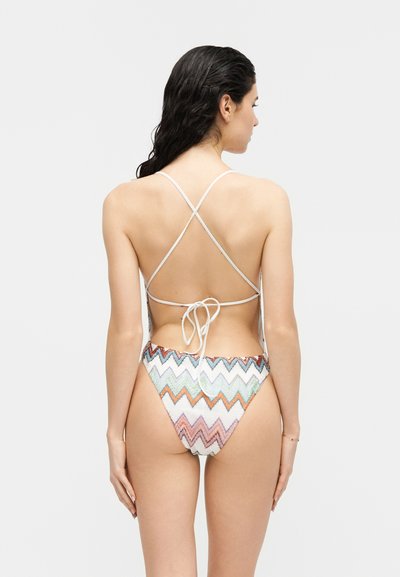 Γυναίκα με βρεγμένα μαλλιά, φορώντας μια ολόσωμη στολή κολύμβησης με πολύχρωμο ζιγκ ζαγκ μοτίβο και δεμένη στο πίσω μέρος, στέκεται με την πλάτη προς το φακό μπροστά σε απλό φόντο.