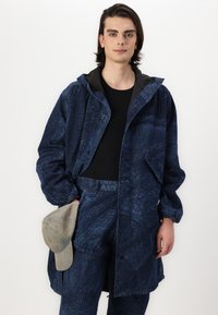 Jūras zila rakstota jakas ar kapuci un pogām, uzvilkta virs melnas krekla. Modelis tur pelēku cepuri. Redzamas teksturētas auduma detaļas.