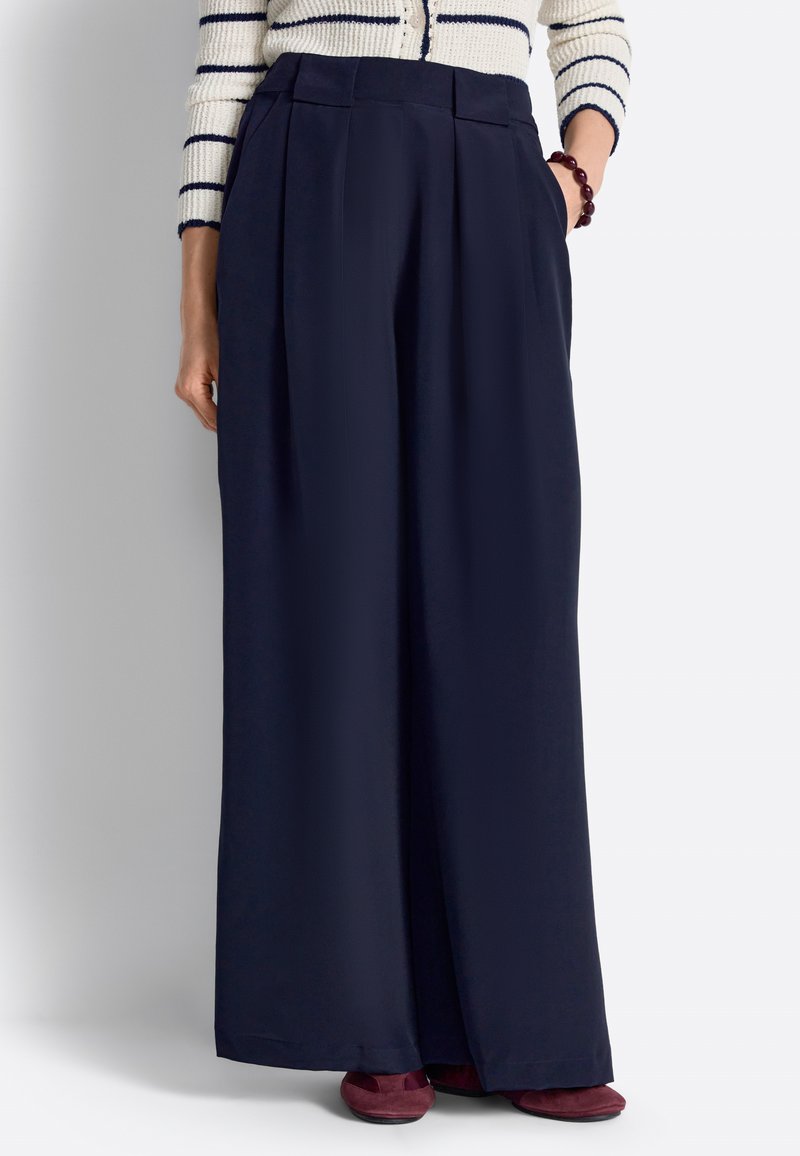 Mujer con pantalones azul marino de talle alto y pierna ancha con pliegues, un suéter a rayas blanco y azul marino, y zapatos planos burdeos, de pie con una mano en el bolsillo.