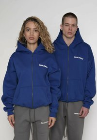 Felpe con cappuccio blu con cerniera, tasca frontale e scritta bianca "MULTIPLY". Abbinate a pantaloni della tuta grigi con un marchio simile. Tessuto morbido al tatto.