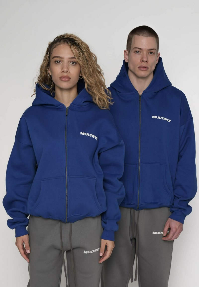 Felpe con cappuccio blu con cerniera, tasca frontale e scritta bianca "MULTIPLY". Abbinate a pantaloni della tuta grigi con un marchio simile. Tessuto morbido al tatto.