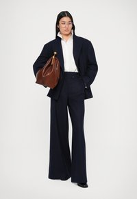 Marinefarbener Oversize-Blazer, weißes Kragenhemd und weit geschnittene Hose. Braune Ledertasche mit goldenen Akzenten. Glatte, strukturierte Texturen.