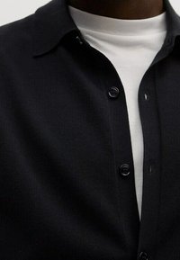 Musta neuletakki pehmeällä tekstuurilla, napitettava edusta ja terävä kaulus valkoisen t-paidan päällä. Ominaisuuksiin kuuluu suuret napit ja istuva malli.