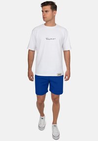 Witte T-shirt met korte mouwen en zwart logo, gecombineerd met blauwe shorts en witte sneaker. De outfit heeft een casual en ontspannen ontwerp.