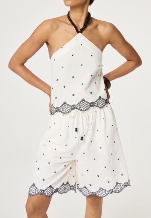 Vrouw draagt witte haltertop en bijpassende short met zwarte kralenketting en zwarte geborduurde geschulpte randen.