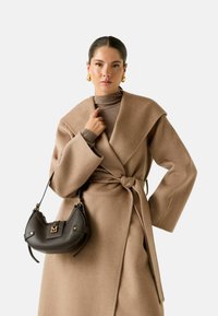 Cappotto di lana beige con colletto ampio e cintura annodata, abbinato a un dolcevita marrone a coste. Una borsa a tracolla in pelle scura completa il look.