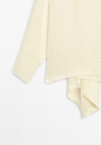 Asymmetrische crèmekleurige top met lange mouwen, zachte textuurstof en een gedrapeerde zoom die een unieke snit en vorm toont.