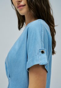 Top azzurro chiaro a maniche corte realizzato in denim con maniche arrotolate. Presenta un dettaglio con bottoni ai polsini e una vestibilità rilassata.