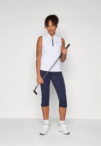 Γυναίκα με λευκή άνευ μανικιών μπλούζα και ναυτικές καπρι παντελόνες, κρατώντας μια ρακέτα γκολφ, να στέκεται μπροστά σε έναν απλό ανοιχτό φόντο.