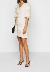 Vestido blanco con escote en V, mangas abullonadas y diseño de capas. Presenta puntos dorados y una silueta fluida. Combinado con chanclas negras.