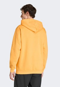 Pomarańczowa bluza z kapturem o miękkiej fakturze, z szerokim dekoltem i ściągaczami przy mankietach. Tył posiada subtelny szczegół w kształcie litery V.