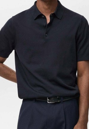 Mann trägt ein dunkelnavyblaues kurzärmeliges Poloshirt, das in passende Hosen mit einem schwarzen Ledergürtel gesteckt ist.