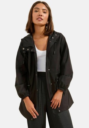 Naine, kes kannab musta kapuutsiga jopet valge soonikkoega tanktopi ja lahtiste mustade pükstega, seisab ühevärvilise tausta ees.