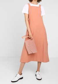 Abito in lino pesco con spalline regolabili, lungo fino al ginocchio, abbinato a una borsa a tracolla in suede rosa chiaro. Il modello indossa scarpe bianche con lacci.